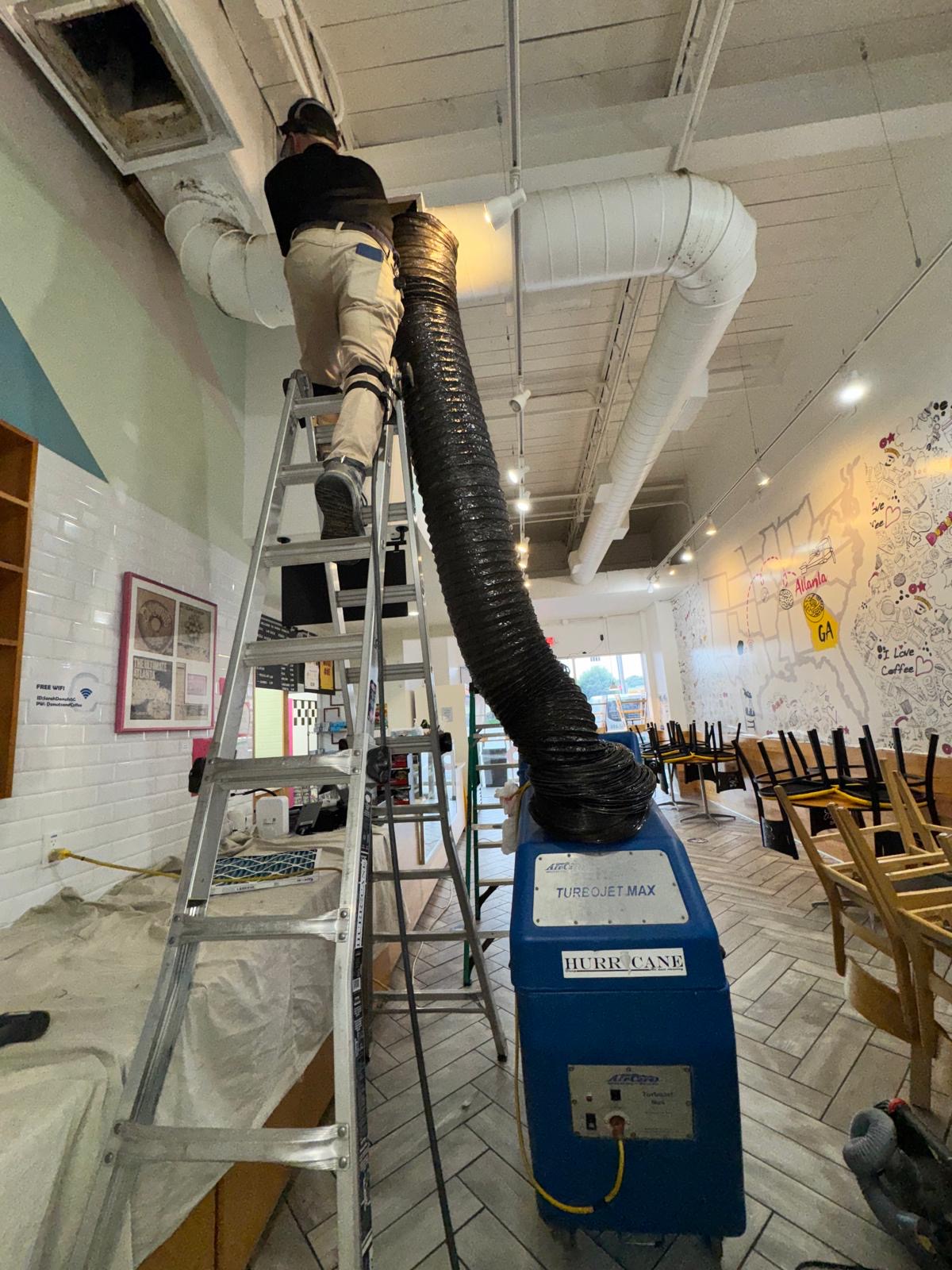 Atlanta coffee shop — technician on ladder cleaning open ceiling supply duct with Hurricane TurboJet Max negative-air machine in the dining room