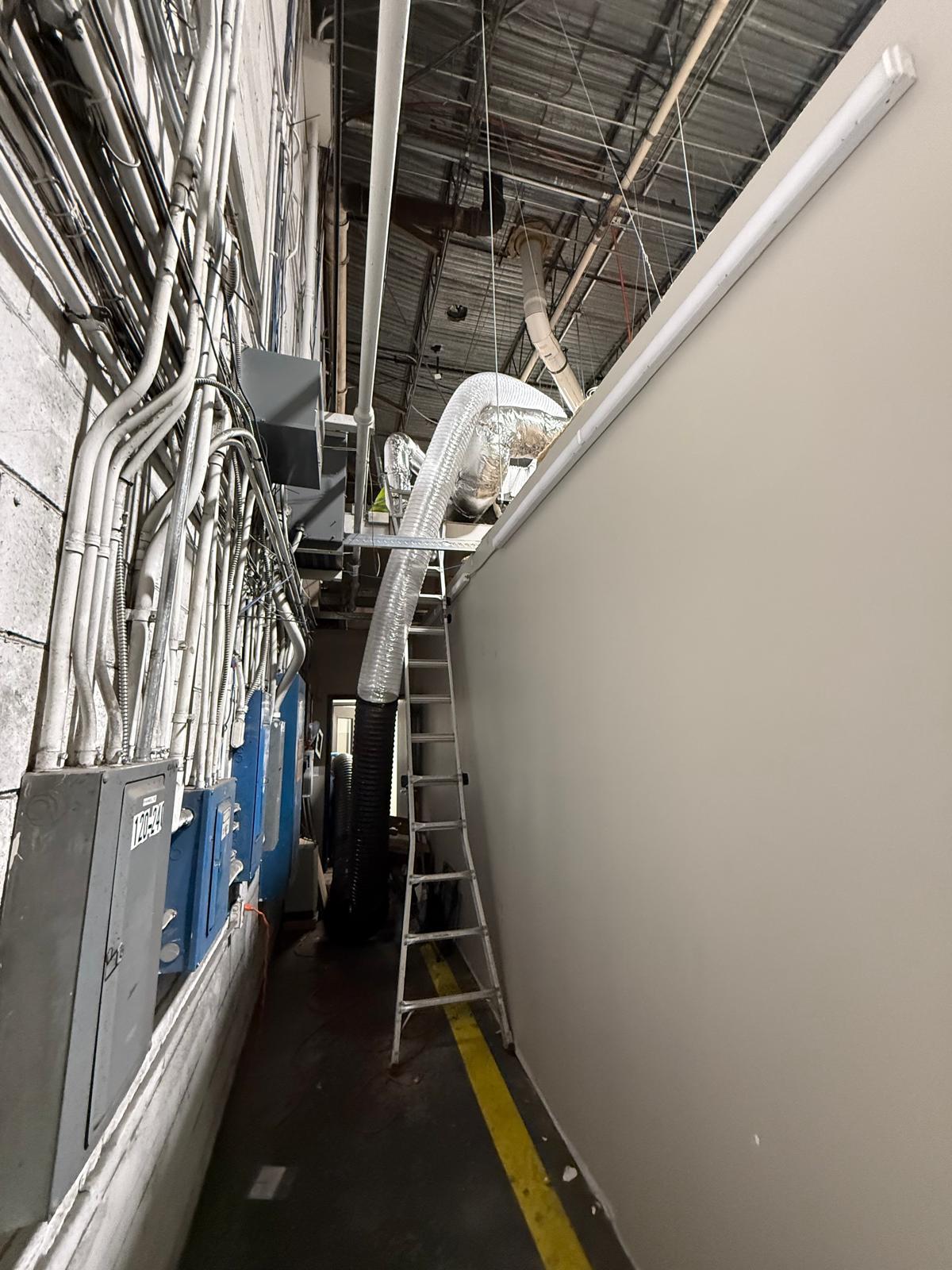 Factory mechanical corridor — flex duct routing past conduit banks and electrical panels